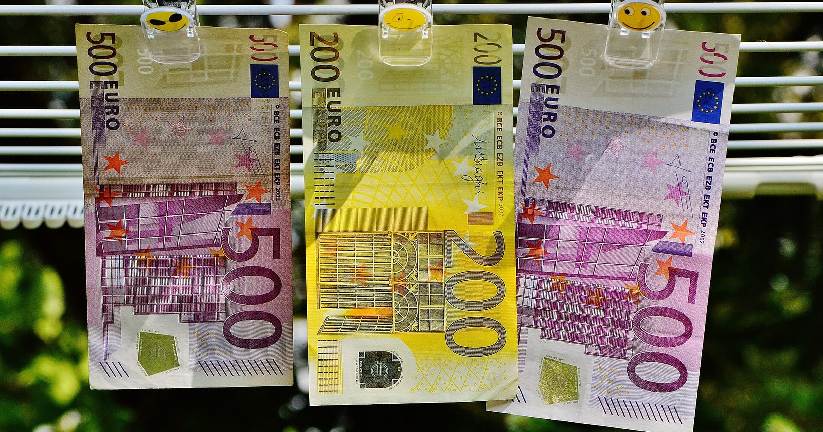 Trzy banknoty euro — dwa po 500 i jeden 200 — przypięte klamerkami do suszarki na tle zieleni